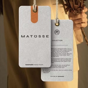 May include: Two rectangular light gray paper hang tags with white string loops. The larger tag features the text MAT&Uuml;SSE and Sustainable Fashion Studio. The smaller tag displays a circular logo, the word COLECTION, and descriptive brand text.