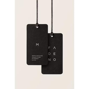 May include: Two rectangular black paper hang tags suspended by thin black strings. The left tag features the letter M and care instructions: Machine wash on cold, Hang dry in shade, Do not bleach, Low iron. The right tag displays the word FADENO.