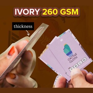 May include: A light purple rectangular clothing tag featuring a green cactus illustration, the text CACTUS CLOTH, and size options S, M, L, and XL. The tag is made of ivory 260 GSM paper, with an arrow pointing to the edge to indicate thickness.