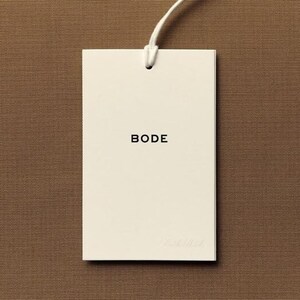 May include: A rectangular off-white paper hang tag featuring the word BODE printed in black capital letters in the center. The tag is attached to a thin white string loop at the top, centered against a solid brown textured background.