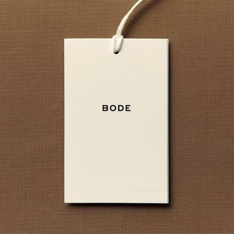 May include: A rectangular off-white paper hang tag featuring the word BODE printed in black capital letters in the center. The tag is attached to a thin white string loop at the top, centered against a solid brown textured background.