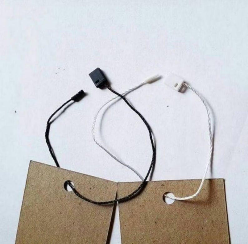 May include: Two rectangular brown kraft paper tags, each featuring a circular hole at the top. One tag is attached to a black string with a black plastic fastener, while the other is attached to a white string with a white plastic fastener.