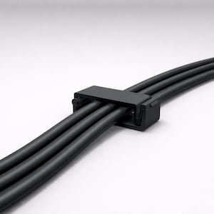May include: A black cable organizer holding three black cables. The organizer is a small, rectangular device with a clip to secure the cables. The cables are curved and appear to be made of a flexible material.