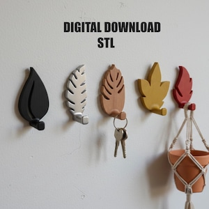 Puede incluir: Ganchos de pared con forma de hojas en negro, plateado, madera, amarillo y rojo. Los ganchos están montados en una pared blanca, con llaves colgadas de un gancho y una pequeña maceta colgante con una maceta de terracota. El texto superior dice "DIGITAL DOWNLOAD STL".