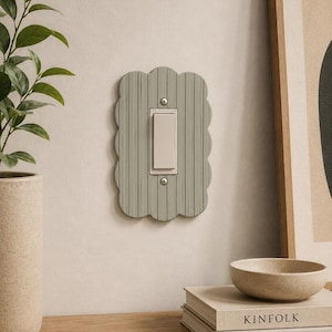 May include: A light green, scalloped-edge light switch cover with a vertical-lined design. The switch is white, and the cover is mounted on a light-colored wall. The image is a close-up of the switch and surrounding decor.