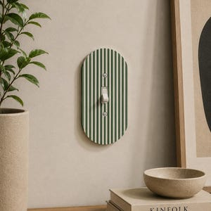 May include: An oval-shaped light switch cover with vertical green and white stripes. The switch is in the on position. The cover is mounted on a light beige wall. A plant in a beige pot is to the left.