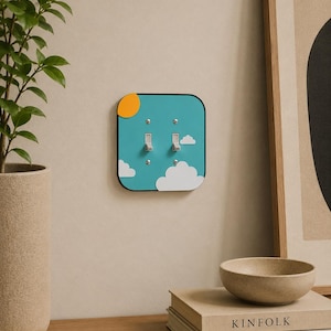 Puede incluir: Una cubierta decorativa para interruptor de luz con un diseño de cielo. La cubierta presenta un fondo turquesa con nubes blancas y un sol amarillo. Se ven dos interruptores basculantes plateados. La cubierta es cuadrada con esquinas redondeadas y un borde negro.