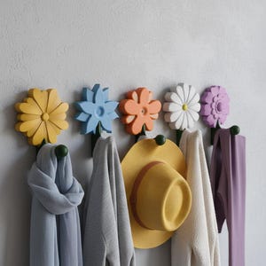 May include: A row of six flower-shaped wall hooks in yellow, blue, orange, white, and purple. Each hook holds a scarf or hat. The hooks are mounted on a light gray wall, offering a decorative and functional storage solution.