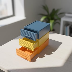 以下が含まれることがあります： 青、黄、オレンジの3色の小さな引き出しが積み重ねられています。各引き出しはわずかに開いており、内部が見えます。引き出しは明るい色の表面に置かれ、背景には窓と植物があります。