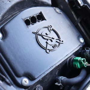 Peut inclure: Une pièce noire de moto avec un emblème circulaire stylisé. La pièce comporte trois ouvertures circulaires et est fixée avec des vis. Des tuyaux et d'autres composants sont visibles.