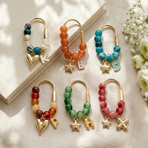 Op de afbeelding: Zes goudkleurige U-vormige karabijnhaak sleutelhangers met kleurrijke kralen in diverse tinten zoals oranje, groenblauw, groen en rood. Elke sleutelhanger is versierd met een goudkleurige metalen bedel in de vorm van een ster of hart en een letter.