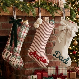 Peut inclure: Trois chaussettes de Noël suspendues à une cheminée. La première est à carreaux verts et rouges avec un nœud en velours vert foncé et le nom Mom en écriture blanche. La deuxième est rayée rouge et blanc avec deux pompons blancs et le nom Annie en écriture rouge. La troisième est crème unie avec un grand nœud crème et le nom Dad en écriture vert foncé.