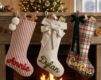 May include: Three Christmas stockings hanging on a mantel. The first is red and white striped with the name Annie in red script. The second is solid cream with a large bow and the name Dylan in green script. The third is red and green plaid with a dark green velvet bow and the name Collins in yellow script.