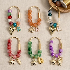 Pode incluir: Seis porta-chaves de metal dourado em forma de U decorados com contas coloridas. Cada porta-chaves apresenta um pendente dourado em forma de estrela, coração ou letra. As letras R, B, K, H e F estão presas a vários aros de contas.