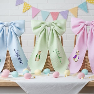 May include: Three pastel-coloured bow-shaped baby bibs with embroidered Peter Rabbit designs and personalised names. The bibs are light blue, green, and pink, displayed in wicker baskets with Easter eggs.