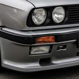 May include: A close-up shot of the front of a silver vintage car. The image focuses on the headlights, indicator, and bumper. The car features two circular headlights, an amber rectangular indicator, and a black grille. The bumper has a curved design.