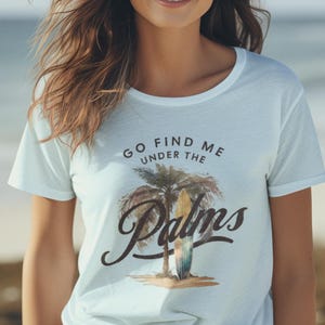 Könnte beinhalten: Hellblaues T-Shirt mit dem Text "GO FIND ME UNDER THE PALMS" und einer Palmen-Grafik. Das T-Shirt wird von einer Person getragen, die an einem Strand mit dem Meer im Hintergrund steht.
