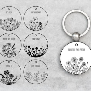 May include: A silver keychain with a round pendant featuring a floral design and the words "BREATHE AND BEGIN." The image also shows six additional round pendants with floral illustrations and motivational phrases like "IT'S OKAY" and "CHASE IT."