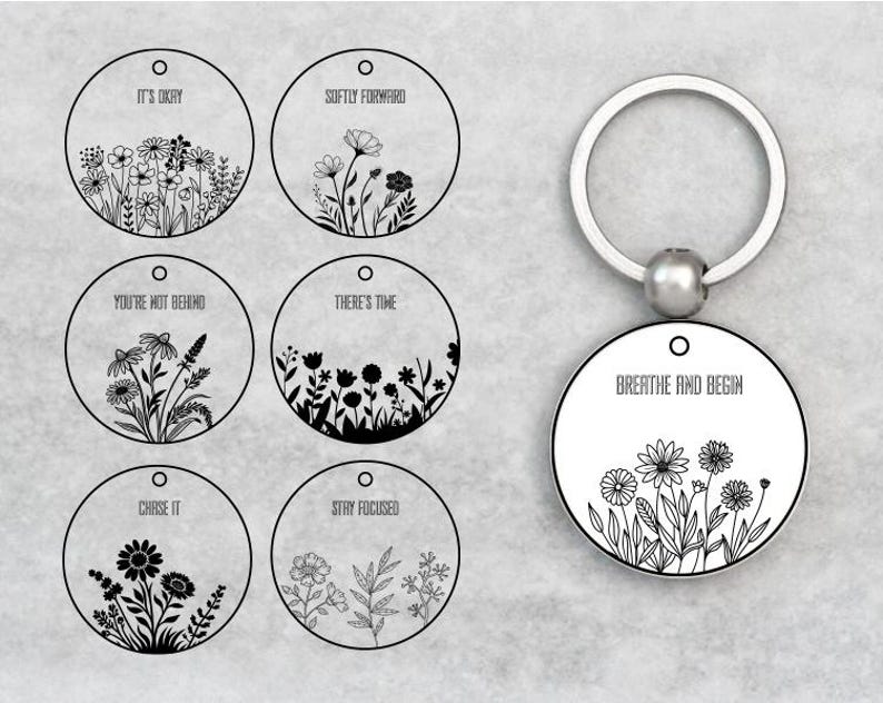 May include: A silver keychain with a round pendant featuring a floral design and the words "BREATHE AND BEGIN." The image also shows six additional round pendants with floral illustrations and motivational phrases like "IT'S OKAY" and "CHASE IT."