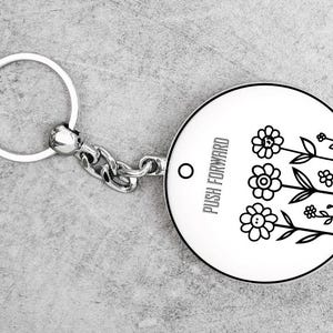 May include: A round, white keychain with a black floral design and the words "PUSH FORWARD." The keychain has a silver chain and ring. The design features three flowers and several leaves.