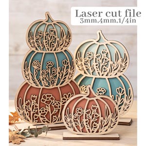 May include: Laser cut file 3mm, 4mm, 1/4in. Three decorative wooden pumpkin displays featuring intricate floral cutouts. One stack of three pumpkins and two individual pumpkins in shades of teal, orange, and natural wood, all on stands.