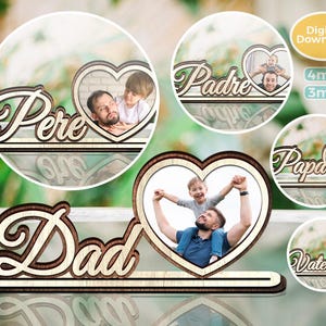 以下が含まれることがあります： 装飾的なフォントで「Dad」という言葉が入った木製のフォトフレーム。フレームには写真を入れるためのハート型の切り抜きがあります。「Pere」、「Padre」、「Papa」、「Vater」という言葉が入った追加のフレームも表示されています。