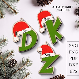 Puede incluir: Adornos navideños de madera con forma de las letras D, K y Z, cada uno pintado de verde y rematado con un gorro de Papá Noel rojo. Los nombres "Daisy", "Kevin" y "Zoey" están impresos en los adornos. El texto "ALL ALPHABET INCLUDED" está en la parte superior.