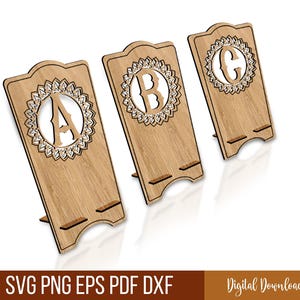 Puede incluir: Tres soportes de teléfono de madera clara, cada uno con una letra diferente (A, B, C) cortada en un diseño circular decorativo. Los soportes tienen una ranura en la parte inferior. También aparece el texto "SVG PNG EPS PDF DXF".