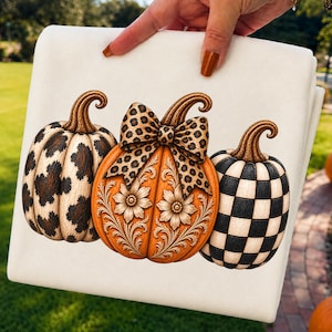 May include: A white fabric item featuring a graphic of three pumpkins. The left pumpkin has a brown leopard print. The center orange pumpkin has a leopard print bow and floral carvings. The right pumpkin features a black and white checkerboard pattern.