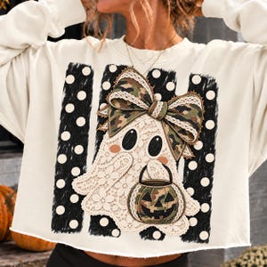 May include: A cream-colored long-sleeve sweatshirt featuring a graphic of a ghost made of white lace. The ghost wears a camouflage bow and carries a camouflage jack-o'-lantern bucket. The design is set against three black vertical stripes with white polka dots.