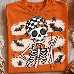 Może przedstawiać: Pomarańczowy t-shirt z grafiką szkieletu w czarno-białej czapce z daszkiem w szachownicę i pomarańczowej muszce. Szkielet wykonuje gest dłoni rock and roll, otoczony czarnymi nietoperzami oraz pomarańczowymi i czarnymi gwiazdami.
