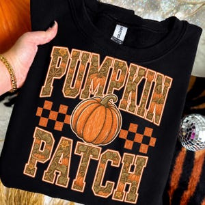 Puede incluir: Una sudadera negra de cuello redondo con el texto PUMPKIN PATCH en letras grandes. Las letras tienen un patrón decorativo de calabazas naranjas y enredaderas verdes. Una calabaza naranja grande se sitúa en el centro con cuadros naranjas.