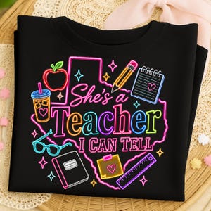 Può includere: Una t-shirt nera con una grafica in stile neon dello stato del Texas. Il design include il testo "She's a Teacher I CAN TELL" circondato da illustrazioni di una mela, una matita, un quaderno, una bevanda, occhiali e un righello.