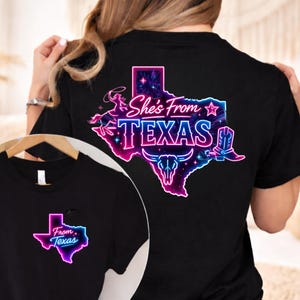 K&ouml;nnte beinhalten: Schwarzes T-Shirt mit einem Neon-Grafikdesign des Bundesstaates Texas und dem Text "She's From Texas". Das Design umfasst einen Cowboy, einen Stern, einen Longhorn-Sch&auml;del und einen Cowboy-Stiefel. Eine kleinere Version des Designs befindet sich auf der Vorderseite.