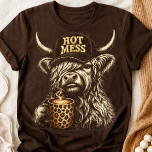 May include: A dark brown t-shirt featuring a graphic of a shaggy Highland cow wearing a black baseball cap with the text "HOT MESS" in gold lettering. The cow is holding a steaming mug with a brown and black leopard print pattern.