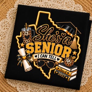 May include: A black t-shirt featuring a gold outline of the state of Texas with the text "She's a SENIOR I CAN TELL". The design includes a leopard print tumbler, a graduation cap, a diploma with a bow, sunglasses, and books labelled "Class of 2027".