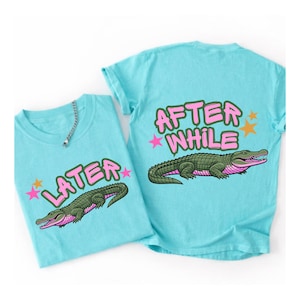 Puede incluir: Dos camisetas de color azul claro con un diseño de cocodrilo de dibujos animados. Una camiseta dice "LATER" y la otra "AFTER WHILE" en letras rosas con burbujas. El cocodrilo es verde con detalles rosas y mira hacia la derecha.