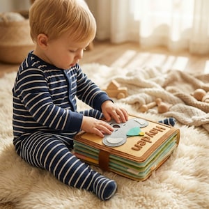 Può includere: Un bambino in una tutina a righe blu navy e bianche è seduto su un soffice tappeto bianco, che interagisce con un libro di feltro. Il libro presenta un design di elefante grigio, il nome "Hayden" e un cinturino in pelle marrone.