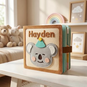 May include: A handmade fabric book with the name "Hayden" on the cover. The cover features a gray koala wearing a party hat. The book has a brown border and a brown strap closure. The pages are visible on the side.