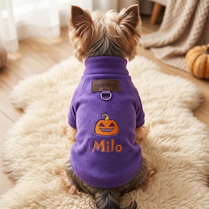 May include: A purple fleece dog sweater featuring an embroidered orange jack-o'-lantern and the name Milo in orange lettering. The garment includes a brown rectangular patch labeled FASHION and a silver metal D-ring attached at the upper back.