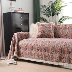 Könnte beinhalten: Ein Sofa mit einem gemusterten Überwurf und passenden Kissen. Der Überwurf und die Kissen weisen ein sich wiederholendes fächerartiges Design in Pfirsich-, Blau- und Brauntönen auf. Der Überwurf hat eine weiße Fransenborte. Ein weißes Kunstfellkissen befindet sich ebenfalls auf dem Sofa.