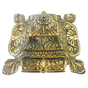 Könnte beinhalten: Verzierter antiker Tintenfass aus Messing mit aufwendigen floralen und scrollartigen Details. Das Tintenfass hat ein quadratisches zentrales Fach mit einem dekorativen Deckel, umgeben von stilisierten Blattmotiven. Das Design ist symmetrisch und detailliert.