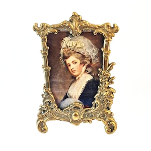 May include: A vintage portrait of a woman in an ornate, gold-coloured frame. The portrait shows a woman wearing a white bonnet and a dark blue dress. The frame has an intricate design with floral and scroll details.