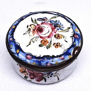 Könnte beinhalten: Eine runde, antike Schmuckdose mit weißem Hintergrund. Der Deckel zeigt ein Blumenmuster mit rosa, roten, blauen und gelben Blumen. Ein blauer Rand mit gelben Punkten umrahmt das Design. Die Dose hat einen Metallrand und -boden.