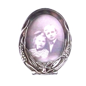 May include: An oval silver-coloured picture frame holding a black and white photograph of two children. The frame features an elaborate floral and leaf design. The frame is standing on a white surface.