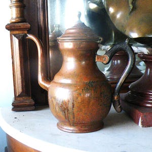 May include: An antique copper pitcher with a curved spout and a dark handle. The pitcher has a lid and is displayed on a marble surface. The pitcher is likely used for serving beverages.