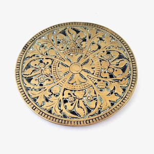 May include: A circular brass medallion featuring an intricate relief pattern of symmetrical scrolling leaves and floral motifs. The design centers on a cross shape surrounded by a circular border, with dark recessed areas highlighting the metalwork.