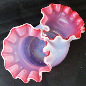 May include: A double-handled, ruffled glass vase with a pink and white iridescent finish. The vase has a scalloped edge and a delicate, feminine design.