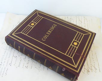 May include: A maroon book with the title "COLERIDGE" in gold lettering and gold decorative detailing. The book is on a sheet of old paper with handwritten text. The cover has a geometric design with floral accents.