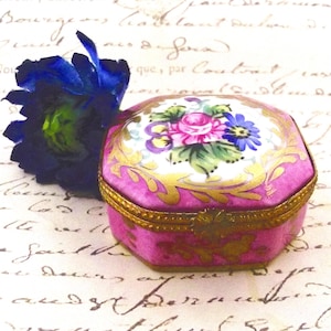 May include: A small, pink, octagonal trinket box with a floral design on the lid. The lid features a painted bouquet of pink roses, blue and purple flowers, and green leaves. Gold trim accents the edges. A blue flower rests beside the box.